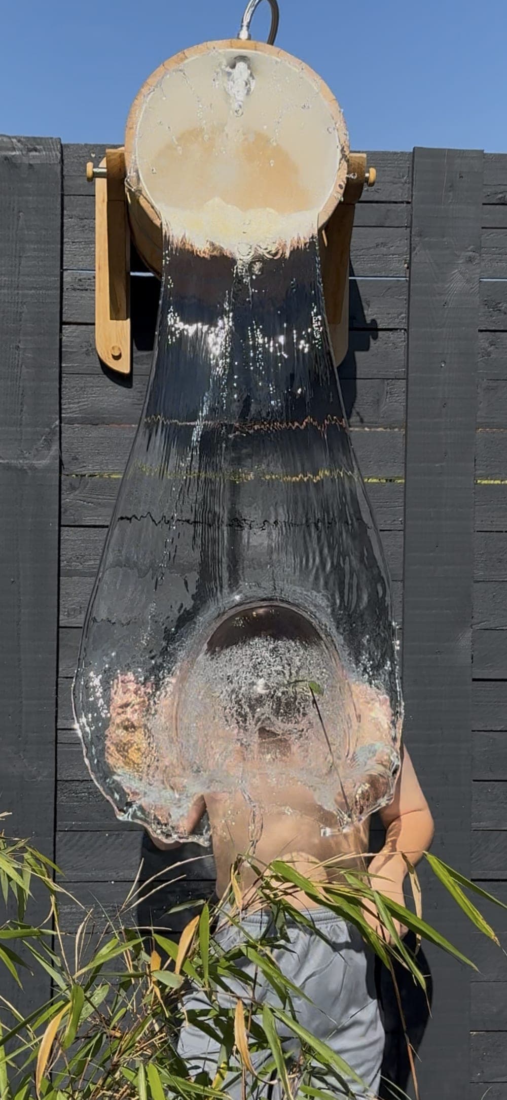 A guest tipping the cedar bucket, cold water sheeting down over them, bamboo in the foreground