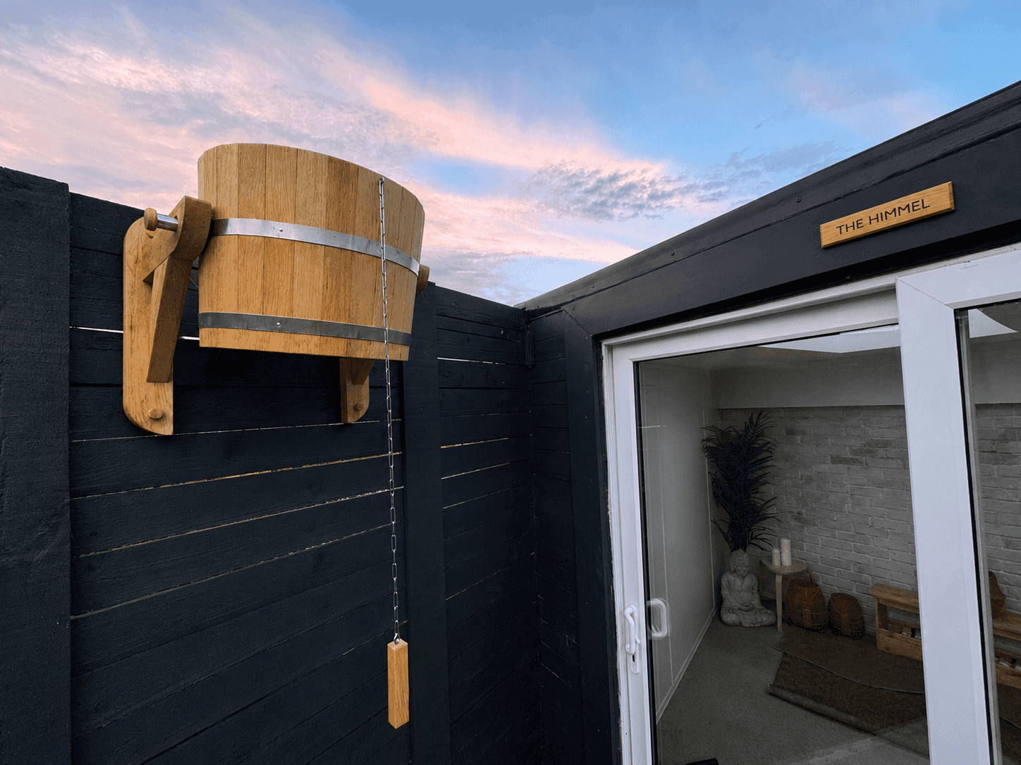 The cedar cold-plunge bucket against the dark cladding at dusk, beside The Himmel